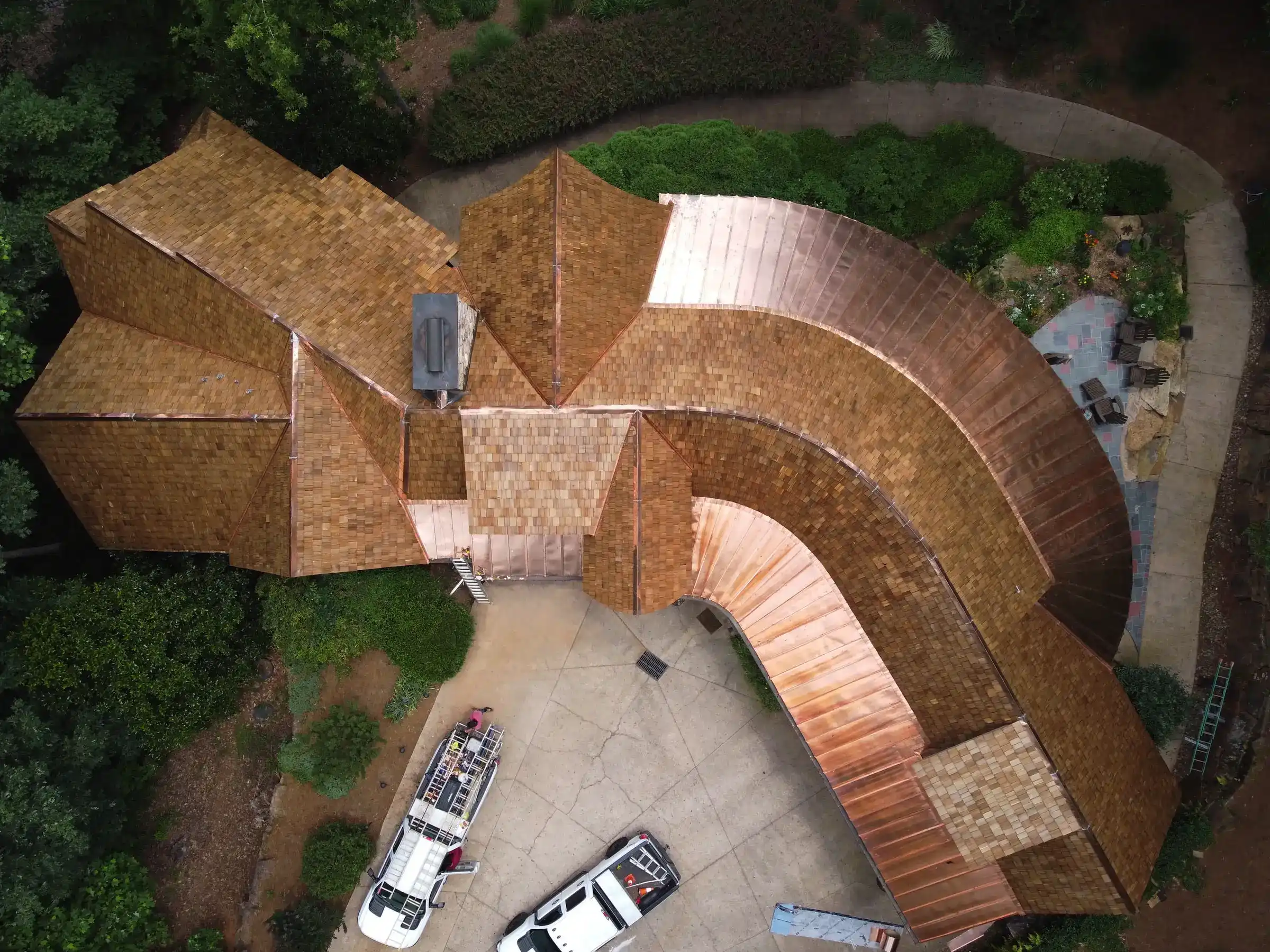 Aerial view of a complex residential roofing project featuring wood shake shingles and curved copper roofing sections, with work trucks and ladders visible in the driveway below.